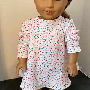 May include: A doll wearing a white dress with pink, blue, and green confetti print. The doll has brown hair and green eyes. The doll is wearing white shoes.