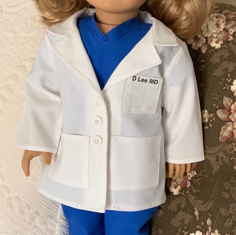Puede incluir: Bata blanca de m&eacute;dico con un top de quir&oacute;fano azul debajo. La bata tiene un bolsillo en el lado izquierdo con una etiqueta de nombre que dice "D Lee MD".