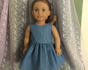 May include: A blue denim dress with a gathered skirt and a sleeveless design. The dress is worn by a doll with brown hair and green eyes.
