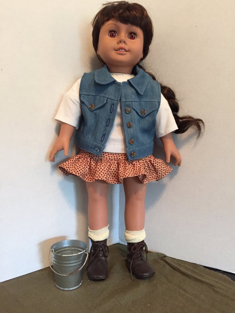 May include: A doll wearing a white shirt, a blue denim vest, a red and white polka dot skirt, white socks, and brown boots. The doll has brown hair and brown eyes. A small silver bucket is in the foreground.