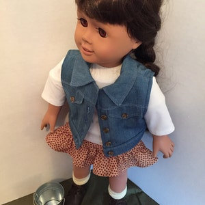 May include: A doll wearing a denim vest, a white shirt, a brown and white patterned skirt, and brown shoes. The doll has brown hair and brown eyes. The doll is standing on a green surface.
