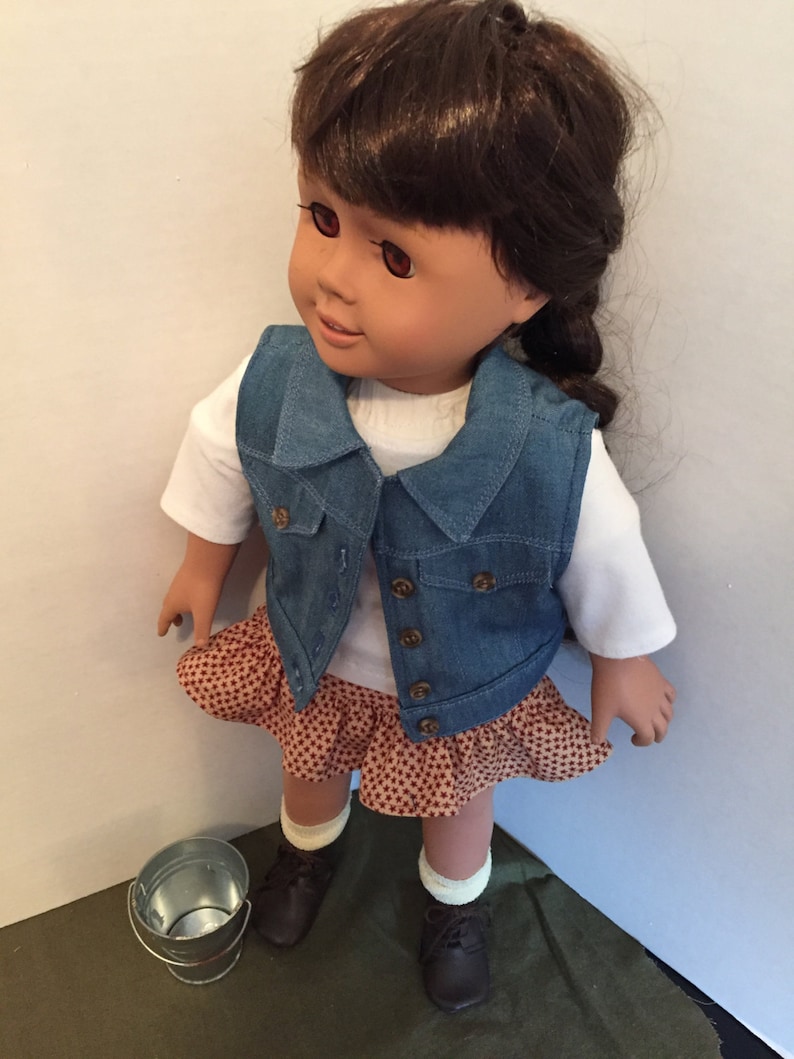 May include: A doll wearing a denim vest, a white shirt, a brown and white patterned skirt, and brown shoes. The doll has brown hair and brown eyes. The doll is standing on a green surface.
