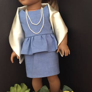 May include: A doll wearing a light blue dress with a white pearl necklace and a cream-colored shawl. The doll is standing in front of a black background with flowers.