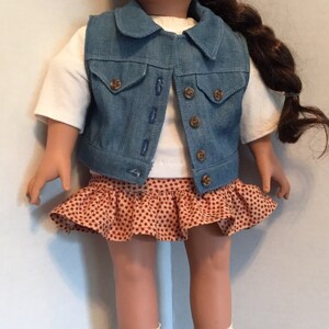 May include: A doll wearing a denim vest, a red and white patterned skirt, white socks, and brown boots. The doll has brown hair and red eyes. A small metal bucket is in the foreground.