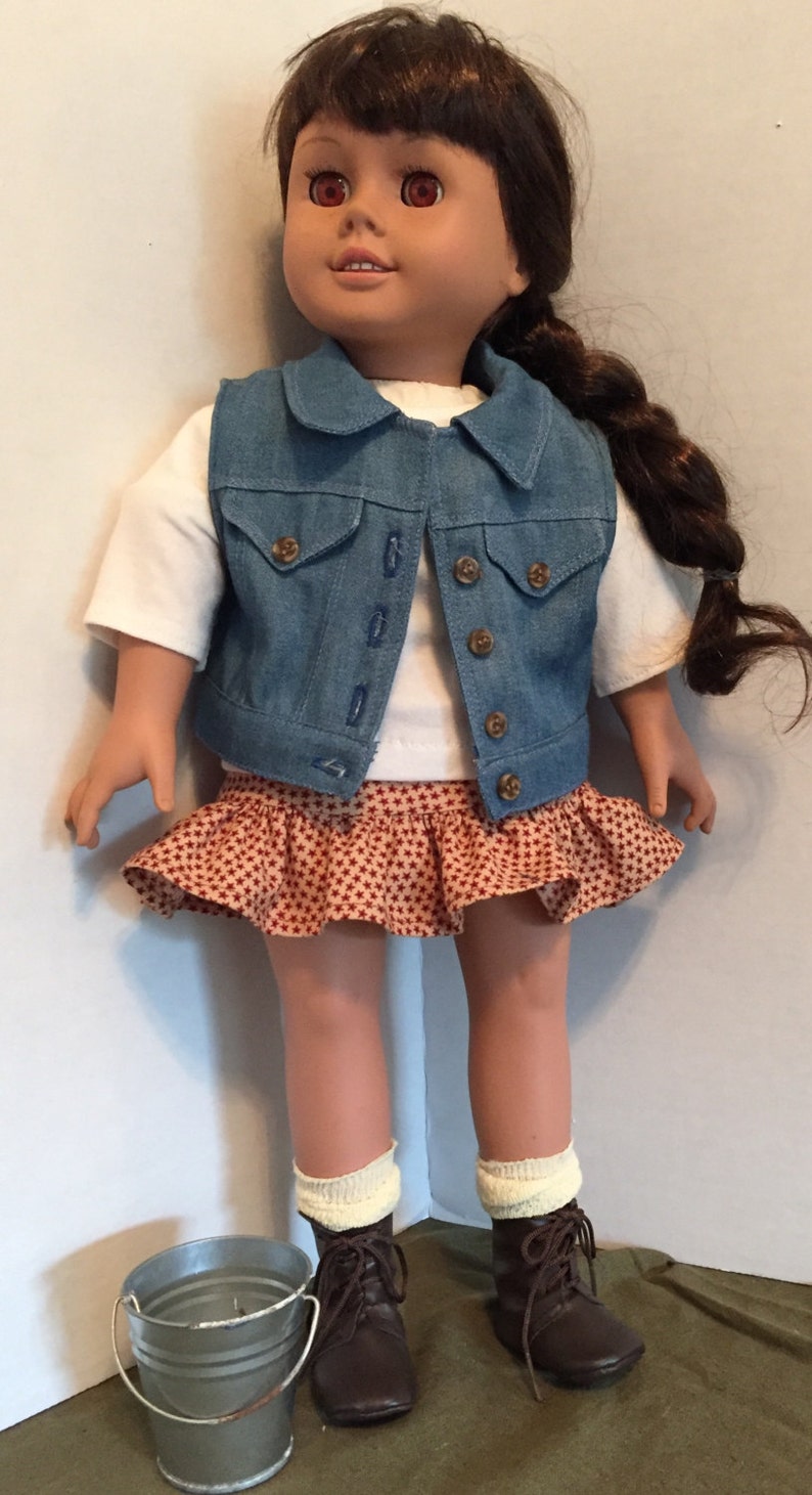 May include: A doll wearing a denim vest, a red and white patterned skirt, white socks, and brown boots. The doll has brown hair and red eyes. A small metal bucket is in the foreground.