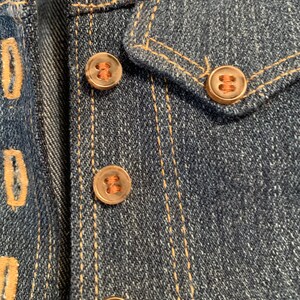 May include: Close-up of a denim jacket with four copper-colored buttons. The buttons have a raised design and are sewn onto the jacket with brown thread.