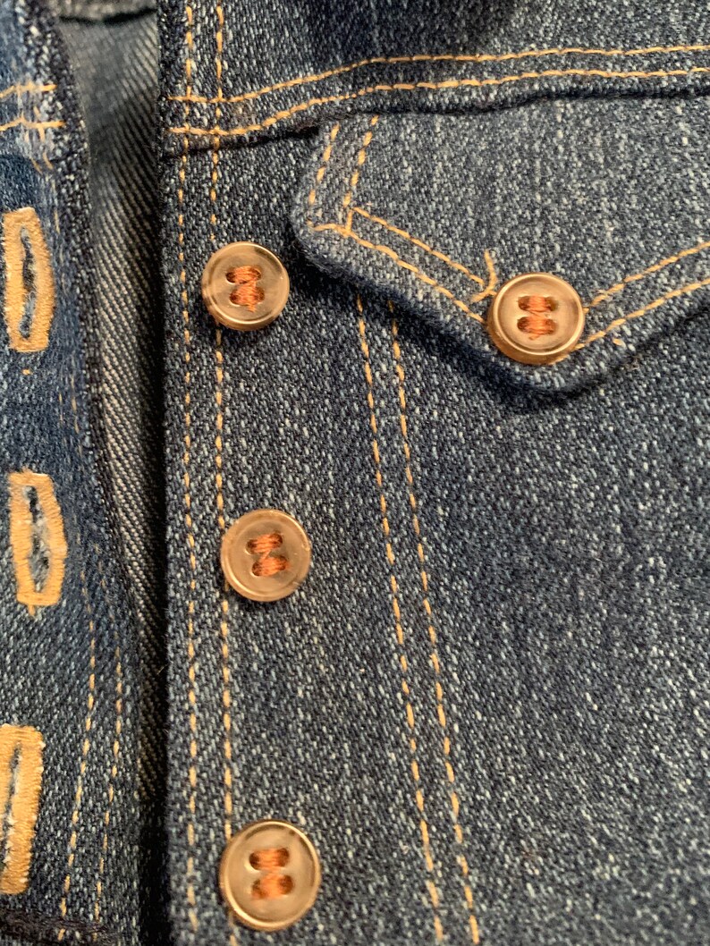 May include: Close-up of a denim jacket with four copper-colored buttons. The buttons have a raised design and are sewn onto the jacket with brown thread.