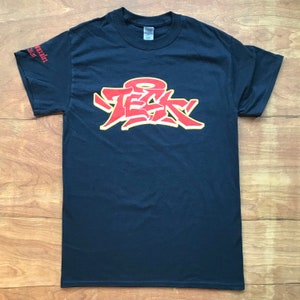 Puede incluir: Camiseta negra con un gráfico rojo y amarillo de la palabra "Tek" en estilo graffiti.