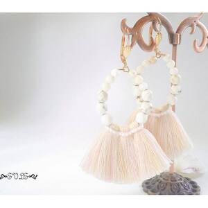May include: A pair of white tassel earrings with white beads and gold accents. The earrings are hanging from a brown metal stand.