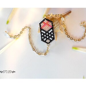 May include: A black and white beaded bracelet with a pink and white geometric charm. The bracelet is on a white surface with a seashell.