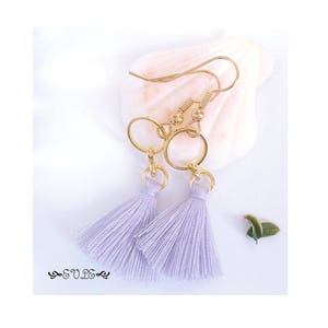 May include: A pair of lavender tassel earrings with gold hoops and a delicate chain. The earrings are hanging from a white background.