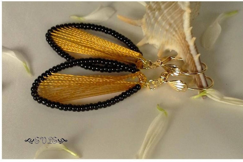 May include: A pair of gold earrings with black beads and yellow thread. The earrings are shaped like a teardrop and have a delicate, feminine design.