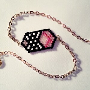 May include: A gold chain bracelet with a black and white beaded charm featuring a pink and white design. The charm is shaped like a rectangle with rounded corners.