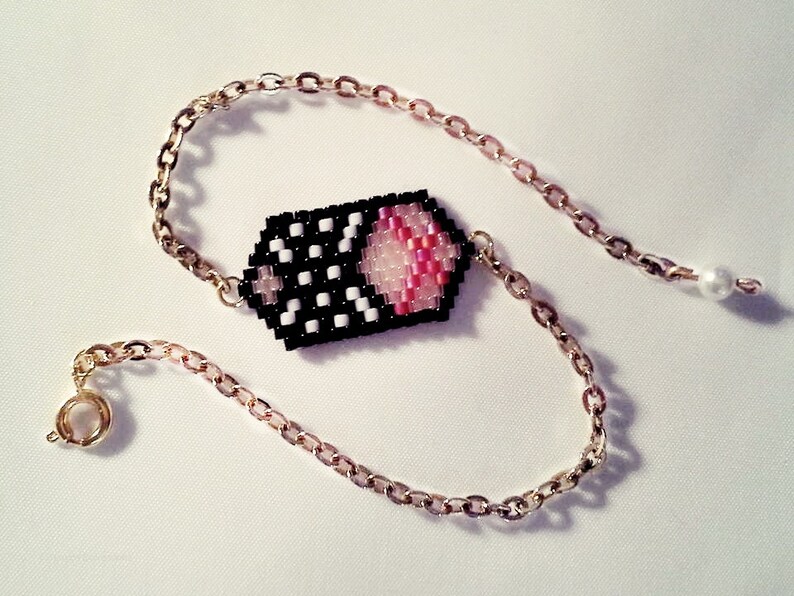 May include: A gold chain bracelet with a black and white beaded charm featuring a pink and white design. The charm is shaped like a rectangle with rounded corners.