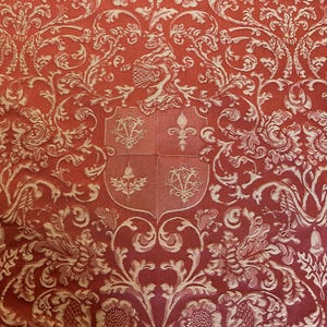 May include: A red fabric featuring a woven damask pattern in a lighter shade. The design includes intricate scrolling acanthus leaves, floral motifs, and a central heraldic shield divided into four quadrants with symbols including a fleur-de-lis.
