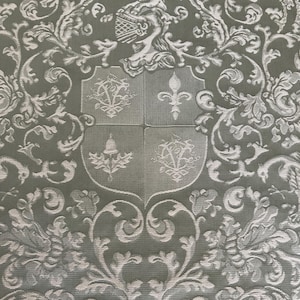 May include: A sage green fabric featuring a woven damask pattern in a lighter shade. The design includes a central shield divided into four quadrants with heraldic symbols, topped by a crest, surrounded by intricate scrolling floral motifs.