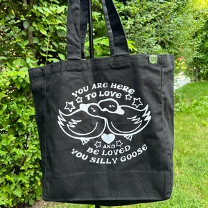 May include: Black tote bag with white text and a graphic of two ducks with a heart. The text reads "You are here to love and be loved you silly goose".