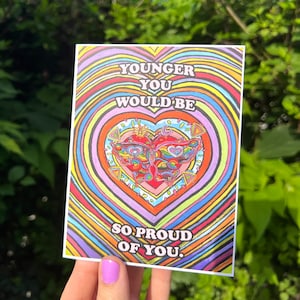 May include: A colorful greeting card with a heart-shaped design and the text "YOUNGER YOU WOULD BE SO PROUD OF YOU." The card features a vibrant butterfly illustration in the center, surrounded by concentric rainbow-colored hearts.