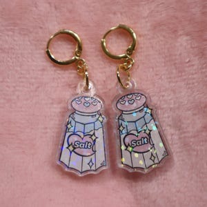 May include: A pair of gold hoop earrings with clear acrylic charms shaped like salt shakers. The salt shakers have a pink top and a white base with a heart that says "Salt" on it. The charms are covered in iridescent glitter.
