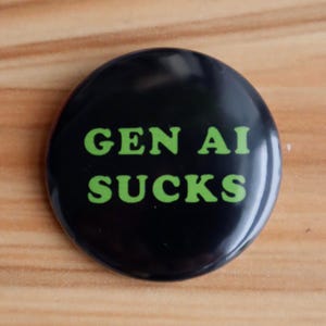 May include: A round, black button with the words "GEN AI SUCKS" printed in bright green, centered on the button. The button is glossy and appears to be made of plastic or metal. The background is a light brown wooden surface.