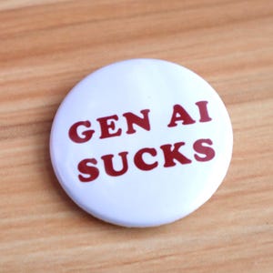 May include: A white button with the text "GEN AI SUCKS" in red letters. The button is round and sits on a wooden surface. The text is centered and clearly visible.