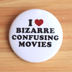 May include: A white button with the text "I ❤️ BIZARRE CONFUSING MOVIES" in black, with a red heart symbol. The button is round and sits on a wooden surface.