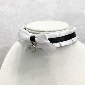 May include: White satin choker with a black ribbon and a silver ring. The choker has a white satin bow with a ruffled edge.
