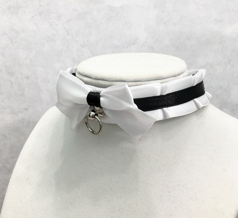 May include: White satin choker with a black ribbon and a silver ring. The choker has a white satin bow with a ruffled edge.