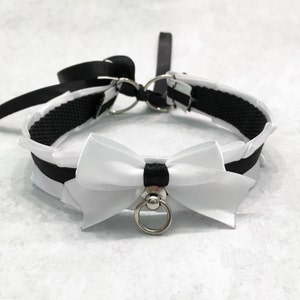 May include: A black and white choker with a white satin bow and a silver ring. The choker is made of a black ribbon and a black elastic band.