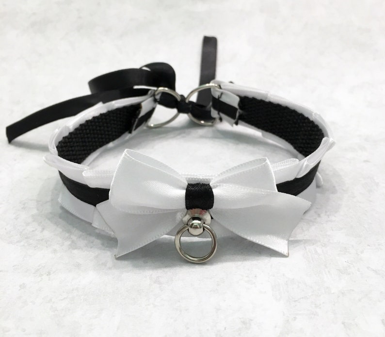 May include: A black and white choker with a white satin bow and a silver ring. The choker is made of a black ribbon and a black elastic band.