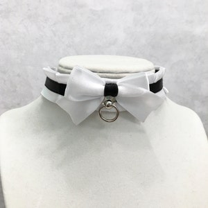 May include: White satin choker with a black ribbon and a silver ring. The choker has a large white satin bow in the center.