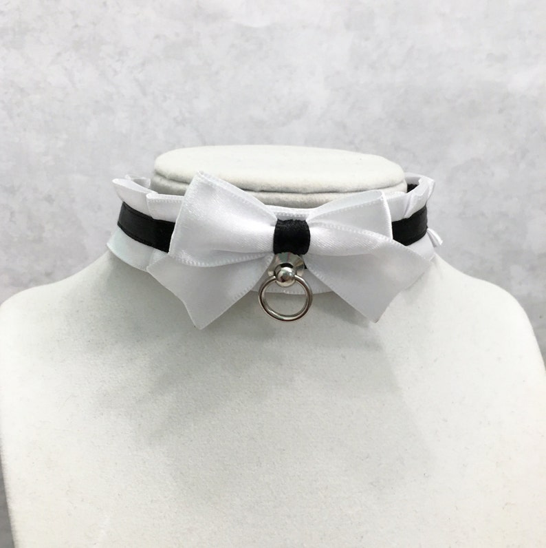 May include: White satin choker with a black ribbon and a silver ring. The choker has a large white satin bow in the center.
