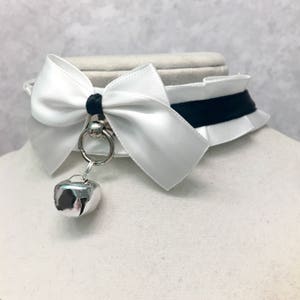 May include: White satin choker with a black ribbon and a white bow. A silver bell hangs from the choker.