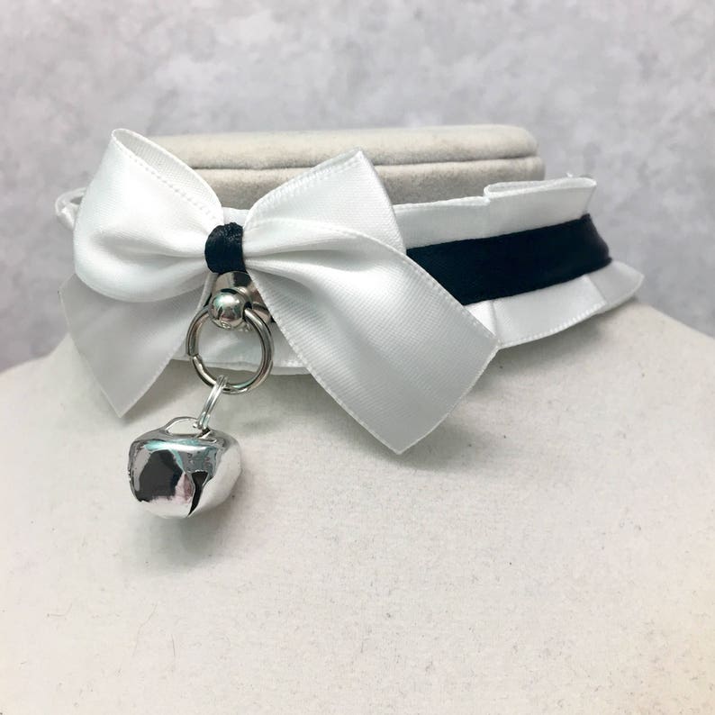 May include: White satin choker with a black ribbon and a white bow. A silver bell hangs from the choker.