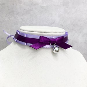 May include: A purple and lavender satin choker with a silver bell charm. The choker has a bow in the front and is tied at the back.