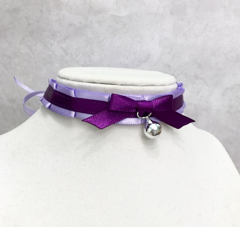 May include: A purple and lavender satin choker with a silver bell charm. The choker has a bow in the front and is tied at the back.