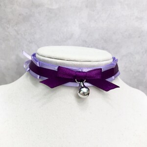 May include: A purple and lavender ribbon choker with a silver bell charm. The choker is tied with a bow in the front.