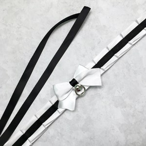 May include: A white satin ribbon choker with a black ribbon trim and a silver ring with a small bell charm. The choker has a large white satin bow.