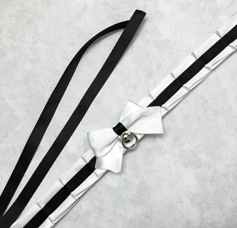 May include: A white satin ribbon choker with a black ribbon trim and a silver ring with a small bell charm. The choker has a large white satin bow.