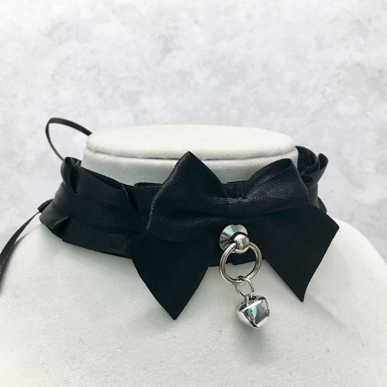 May include: A black satin choker with a large bow and a silver bell charm. The choker has a ruffled edge and a silver ring.