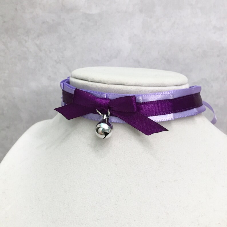 May include: A purple and lavender satin choker with a silver bell charm. The choker has a bow in the front.