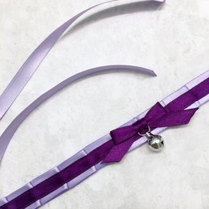 May include: A purple and lavender satin choker with a bow and a silver bell charm.