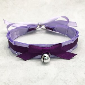 May include: A lavender satin choker with a purple satin bow and a silver bell. The choker is tied with a bow at the back.