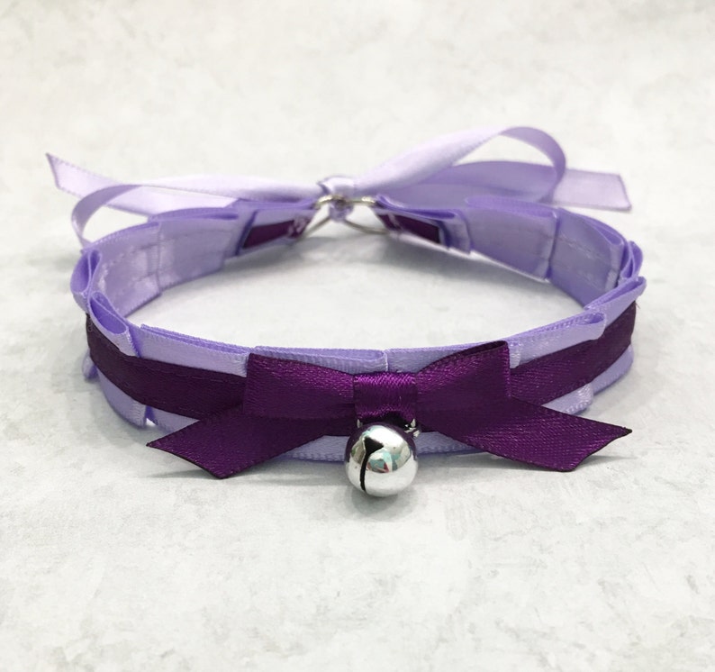 May include: A lavender satin choker with a purple satin bow and a silver bell. The choker is tied with a bow at the back.