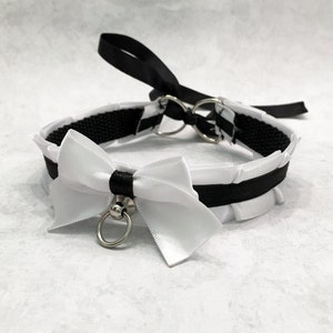 May include: A black and white choker with a white satin bow and a silver ring. The choker is made of black ribbon and has a ruffled edge.