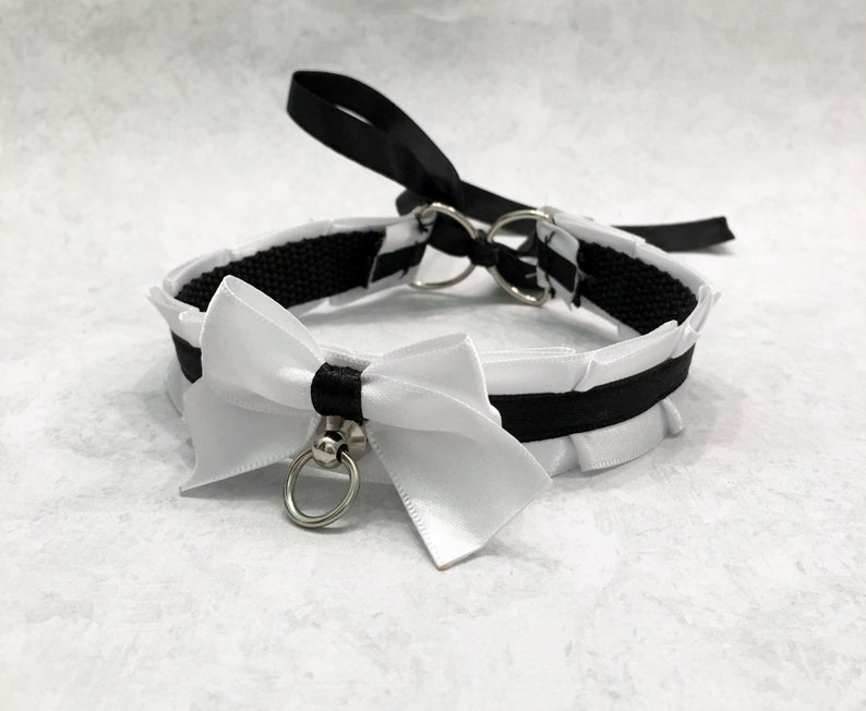 May include: A black and white choker with a white satin bow and a silver ring. The choker is made of black ribbon and has a ruffled edge.