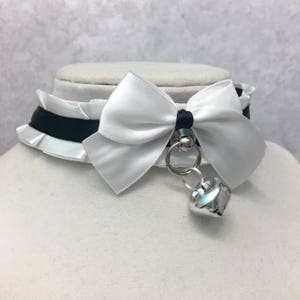 May include: A white satin choker with a black ribbon and a white bow. The choker has a silver heart-shaped charm attached to it.