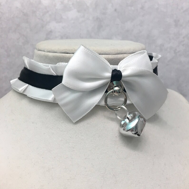 May include: A white satin choker with a black ribbon and a white bow. The choker has a silver heart-shaped charm attached to it.