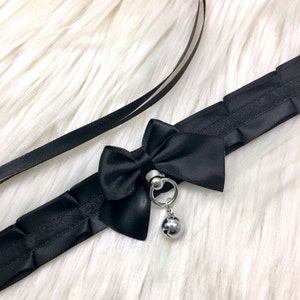 May include: Black satin choker with a large black bow and a silver bell charm. The choker has a silver ring with a metal ring attached to the bell.