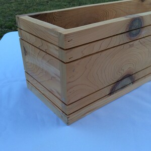 May include: A rectangular wooden planter box with a natural wood finish. The planter has a simple design with three horizontal slats on the front and sides.